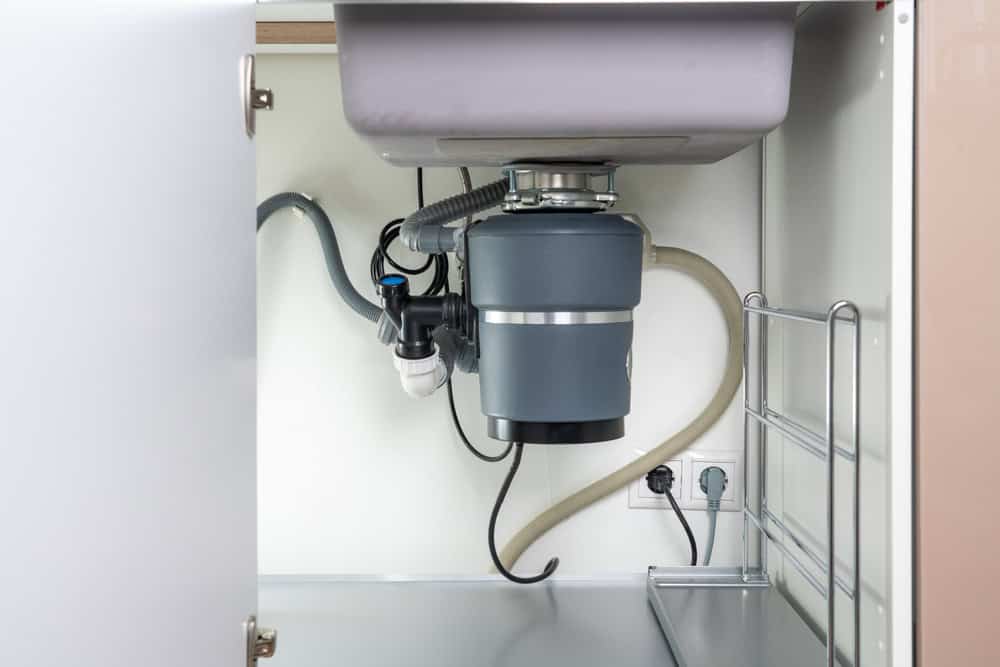 Open cabinet showcasing an installed garbage disposal unit under a sink, expertly connected to plumbing. A power outlet with plugged-in cords is visible in the background. Nearby, a metal rack is attached inside the cabinet. For installations like this, rely on top-notch plumbing services in Cook County, IL.