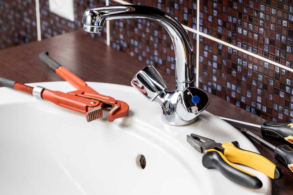 A close-up of a bathroom sink with a shiny silver faucet showcases various plumbing tools, including a pipe wrench and pliers, on the sink and wooden countertop. The dark mosaic tile background highlights the expertise of an emergency plumber in Cook County, IL.