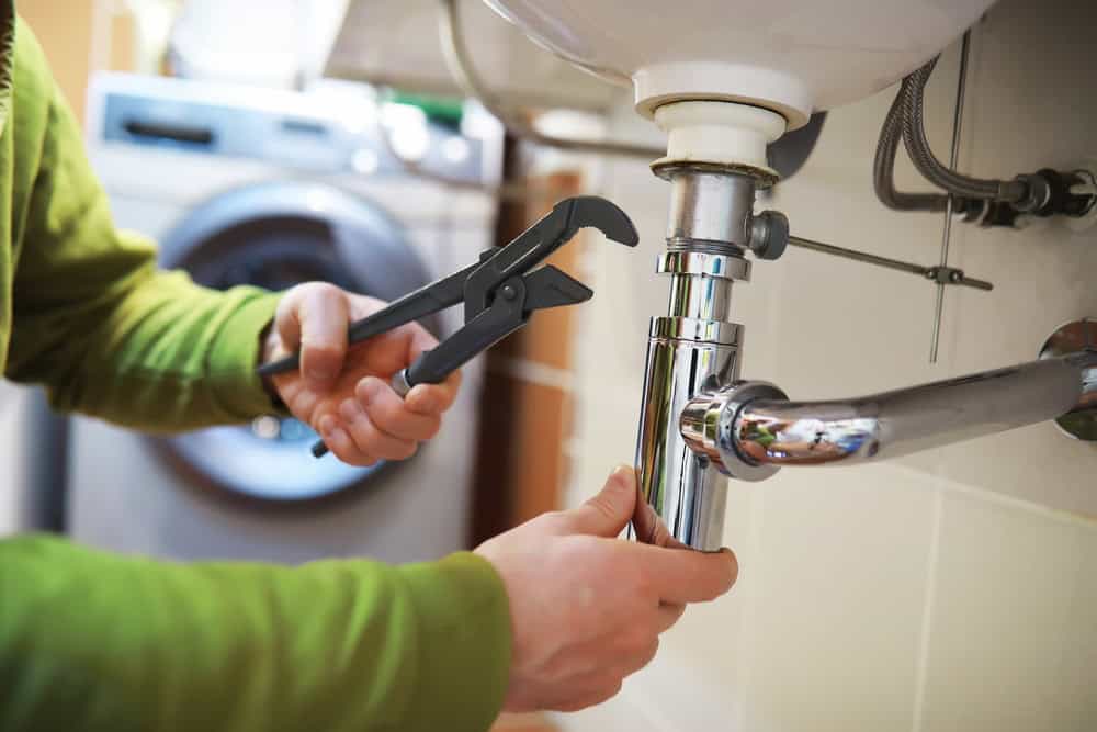 A person in a green shirt skillfully uses a wrench to adjust the pipes under a sink, embodying the precision of an emergency plumber in Cook County IL. In the background, a washing machine sits ready for action.