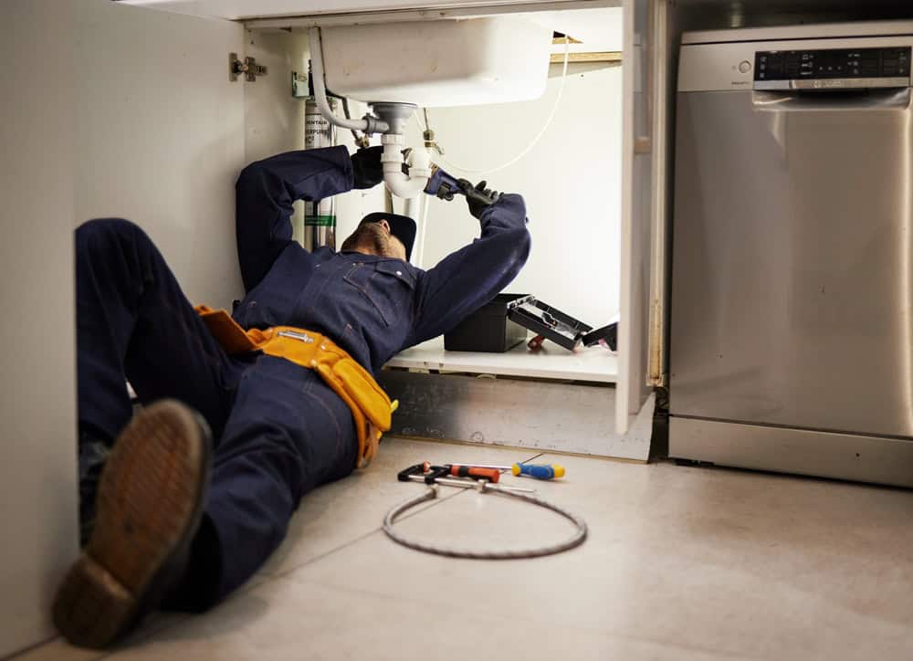 A person in a blue jumpsuit and yellow tool belt lies on their back under a kitchen sink, providing emergency plumbing services in Cook County, IL. Using a wrench on the plumbing, they are surrounded by tools and a toolbox scattered on the floor, with a dishwasher visible beside them.