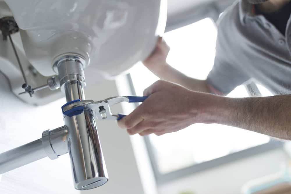 A person uses a wrench to tighten or loosen the pipe under a sink, with natural light streaming through a nearby window. The focus on hands and tools highlights plumbing services Cook County IL, indicating a repair or installation task.
