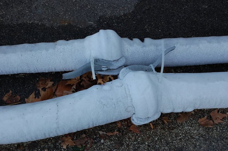 Two large, frost-covered pipes with metal straps lie parallel against a dark textured ground. Fallen brown leaves are scattered around the pipes, contrasting with the white frost.