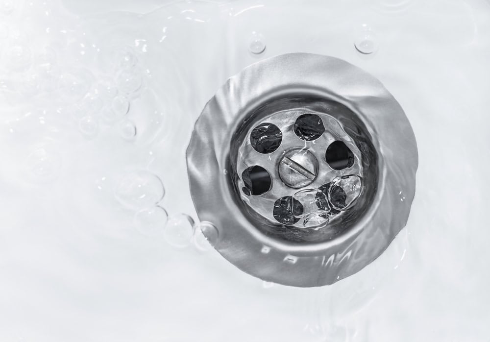 Close-up of a metal drain in a sink with water flowing around it. Water bubbles are visible, creating a swirling effect in the clear liquid as it moves towards the drain. The image captures the shiny, reflective surface of the drain.