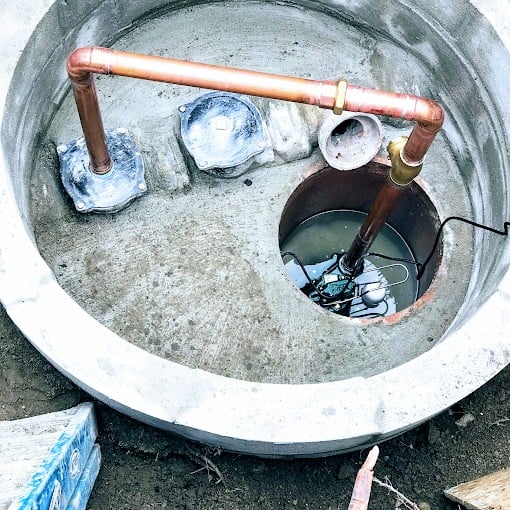 Concrete water well with copper piping and open cover. Inside, there's visible water with submerged equipment. The area is surrounded by dirt, and a wooden plank is nearby, suggesting ongoing work.