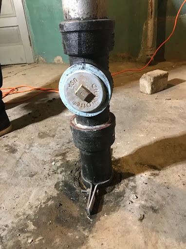 A close-up of a vertical metal sewer pipe with a cleanout cap, set in a concrete floor. The surrounding floor shows signs of water staining or leakage, possibly needing a plumber Chicago & Cook County, IL. An orange extension cord and a concrete block are visible in the background.