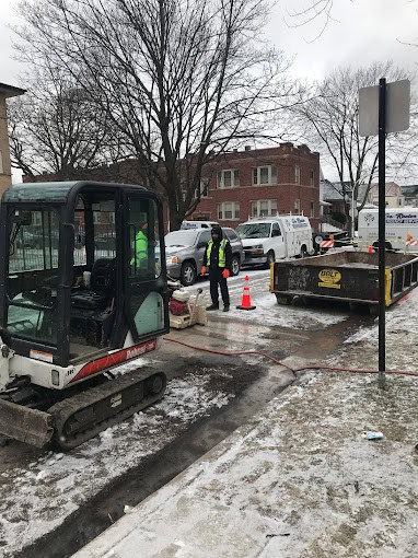 A construction crew works on a sidewalk in a residential area. Equipment includes a mini excavator and an open trench. Orange cones mark the site. Nearby are utility trucks. Snow covers the ground and trees are in the background.