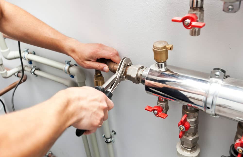 Close-up of a person using a wrench to tighten a valve on a shiny metal pipe. Surrounding are several connected pipes and red-handled valves against a white wall, indicative of plumbing work.