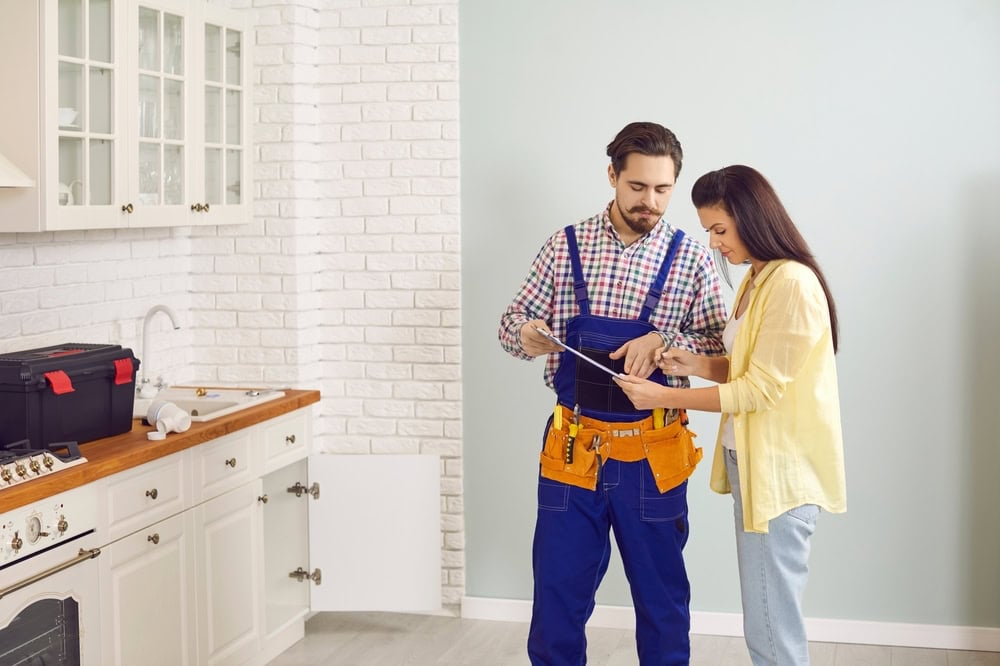 A handyman in a blue jumpsuit with a tool belt and a woman in casual clothes look at a clipboard together in a modern kitchen, discussing repairs. A toolbox and cabinet doors are visible nearby.