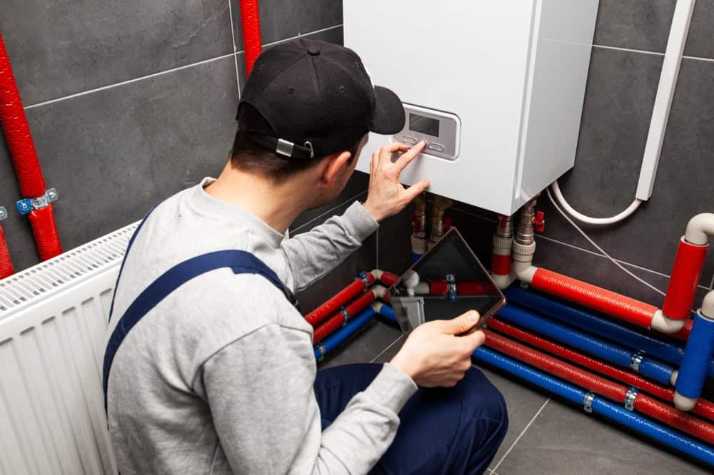 A worker in a cap and overalls checks the control panel of a wall-mounted boiler in a utility room, holding a digital tablet, with red and blue pipes visible on the floor and walls.