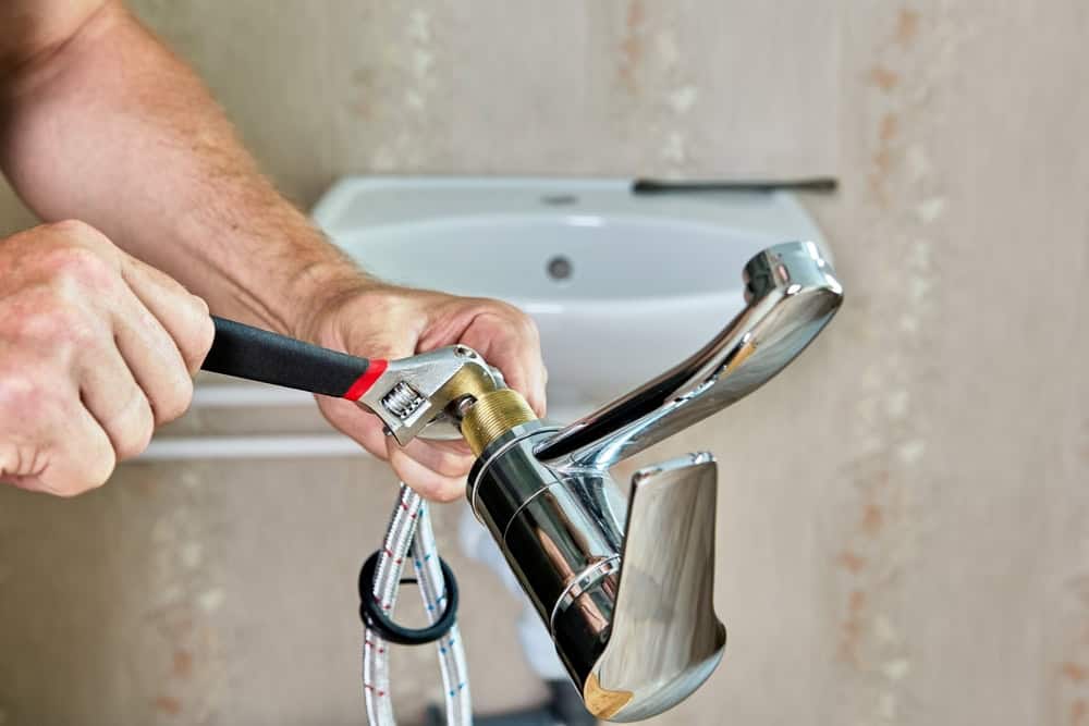 A person uses a wrench to tighten a chrome bathroom faucet. The faucet has attached hoses and is being installed in front of a blurred sink and wall background.