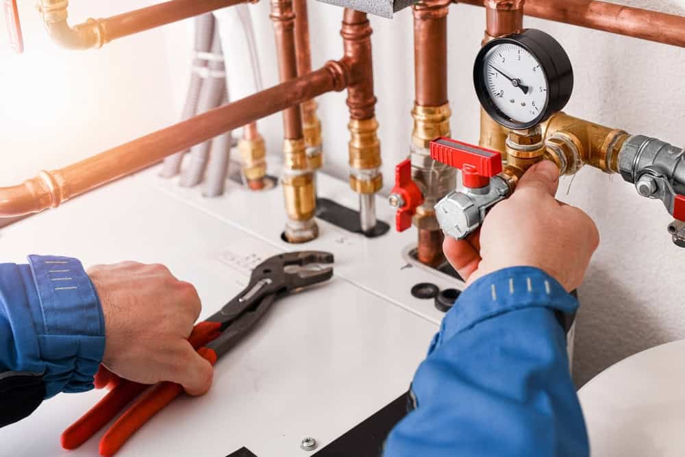 A person wearing a blue work jacket is adjusting a plumbing valve with a wrench. Various copper pipes and gauges are visible, indicating maintenance or repair work in progress on the plumbing system.