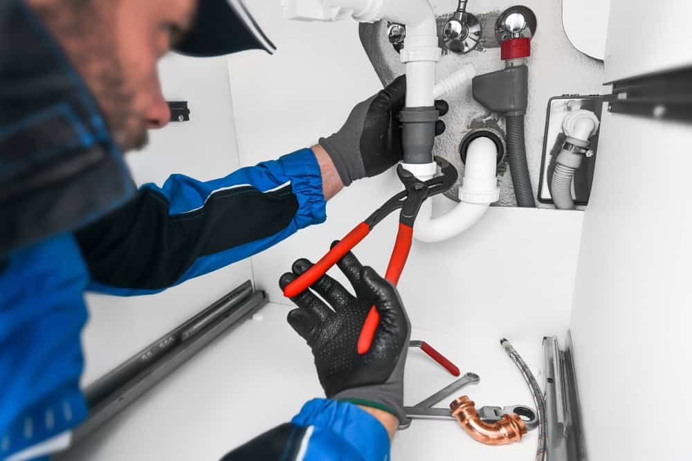 A plumber wearing gloves uses a pipe wrench to tighten a white pipe under a sink. Various plumbing tools and pipes are visible on the floor of the cabinet.