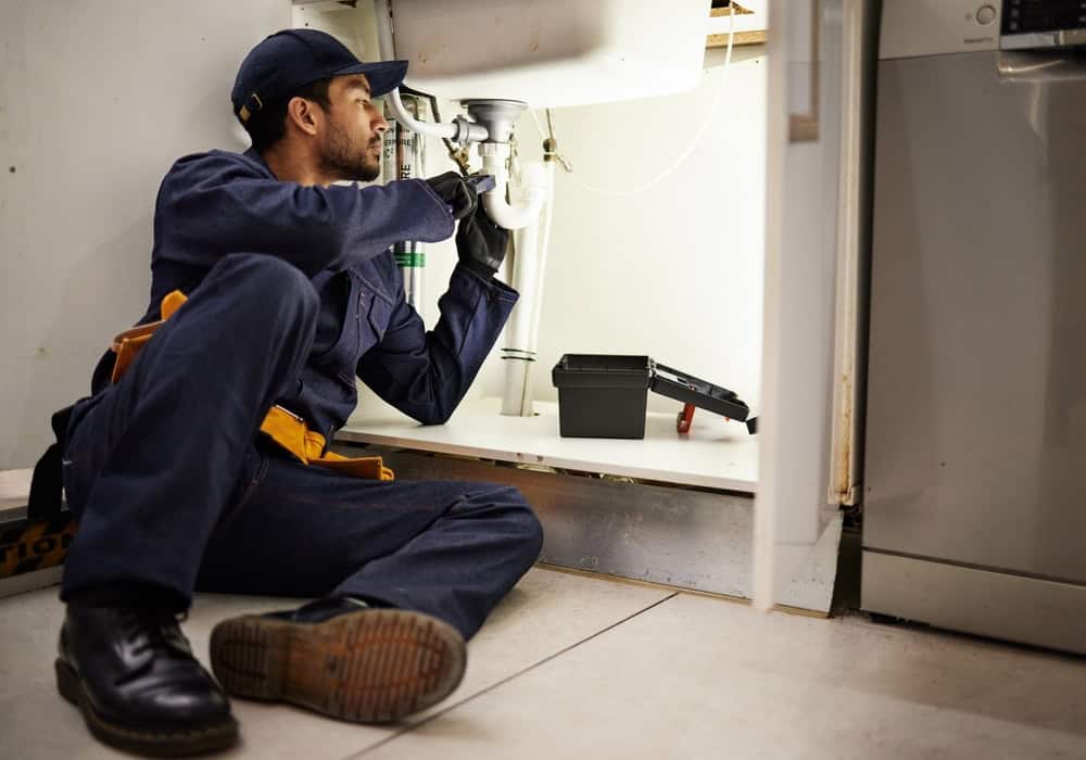 A plumber in a blue uniform is sitting on the floor under a sink, fixing pipes. He is holding tools and looking focused. A toolbox is open beside him on the floor, and a dishwasher is visible in the background.
