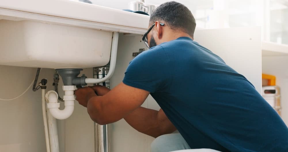 A person wearing glasses and a blue shirt is fixing pipes under a kitchen sink, working with plumbing tools in a bright, modern room.