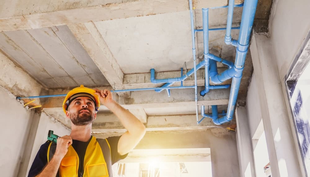 A construction worker wearing a yellow hard hat and vest examines blue water pipes installed on a building ceiling, holding a wrench and looking up thoughtfully. Sunlight shines through a window.