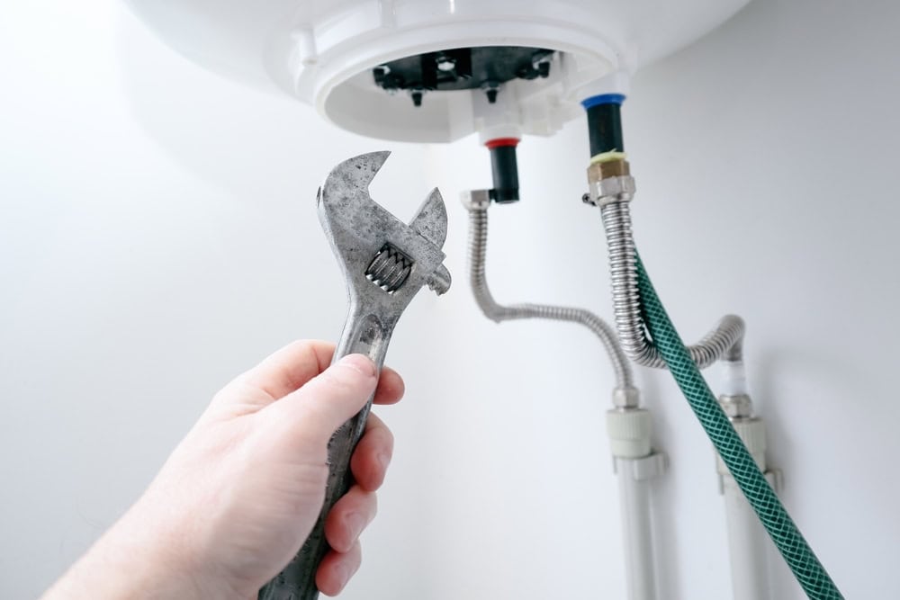 Hand holding an adjustable wrench beneath a white water heater, focusing on the plumbing connections. The image shows metal pipes, a flexible hose, and valve attachments marked with red and blue indicators for hot and cold water.