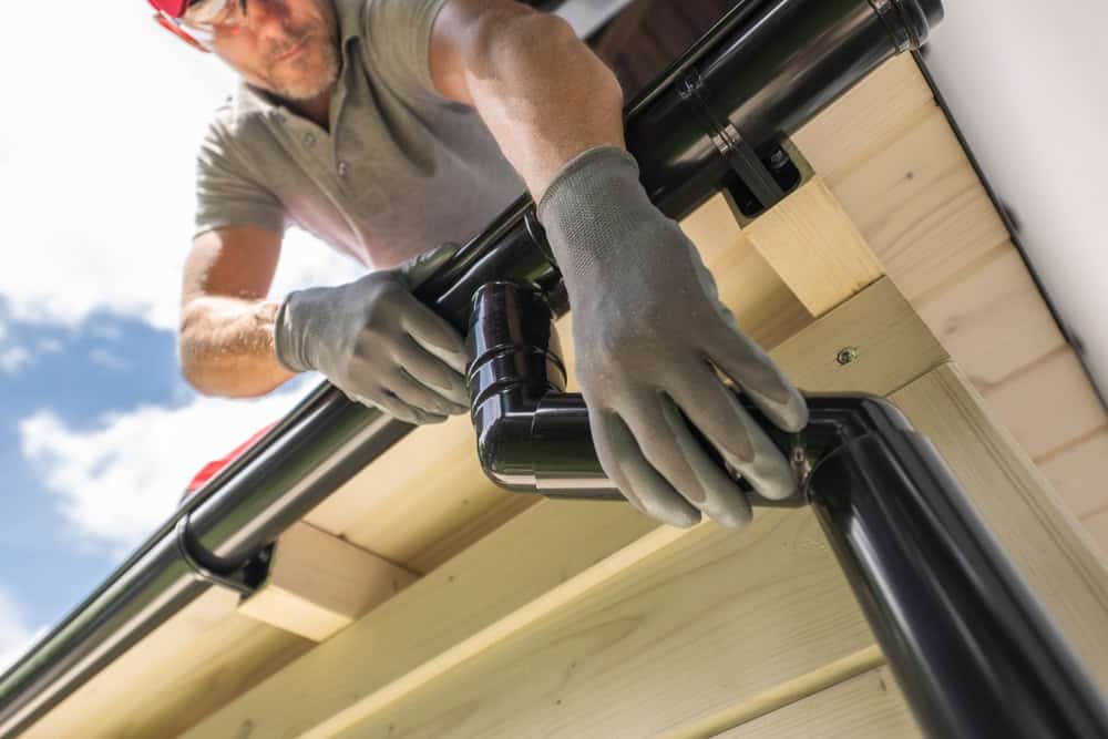 A person wearing gloves installs a black rain gutter system on the edge of a wooden roof, securing a corner joint on a sunny day.