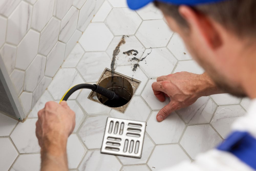 A plumber uses a drain snake to clear a clogged drain in a tiled bathroom floor. The drain cover is removed and set nearby. The plumber's hands and tools are visible close to the drain opening.