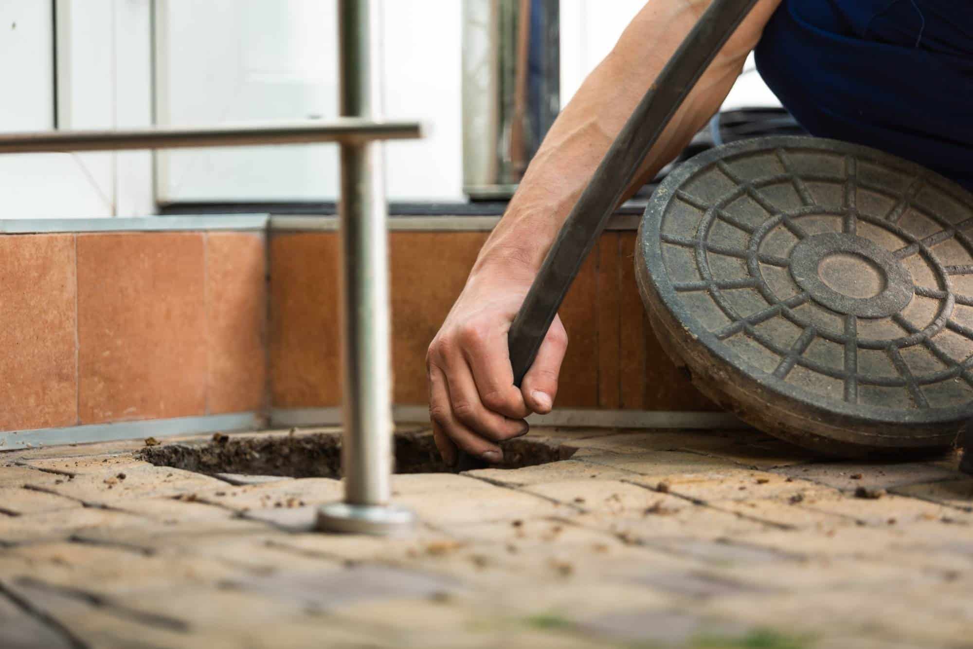 A person’s hand lifts a round manhole cover and inserts a hose into an opening in a tiled floor, suggesting maintenance or cleaning work is being done on a drain or sewer system.