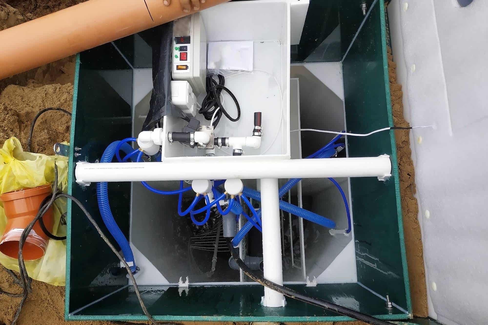 An overhead view of a wastewater treatment unit with control panel, pipes, blue hoses, and electronic components, installed in a green rectangular container partially embedded in the ground.