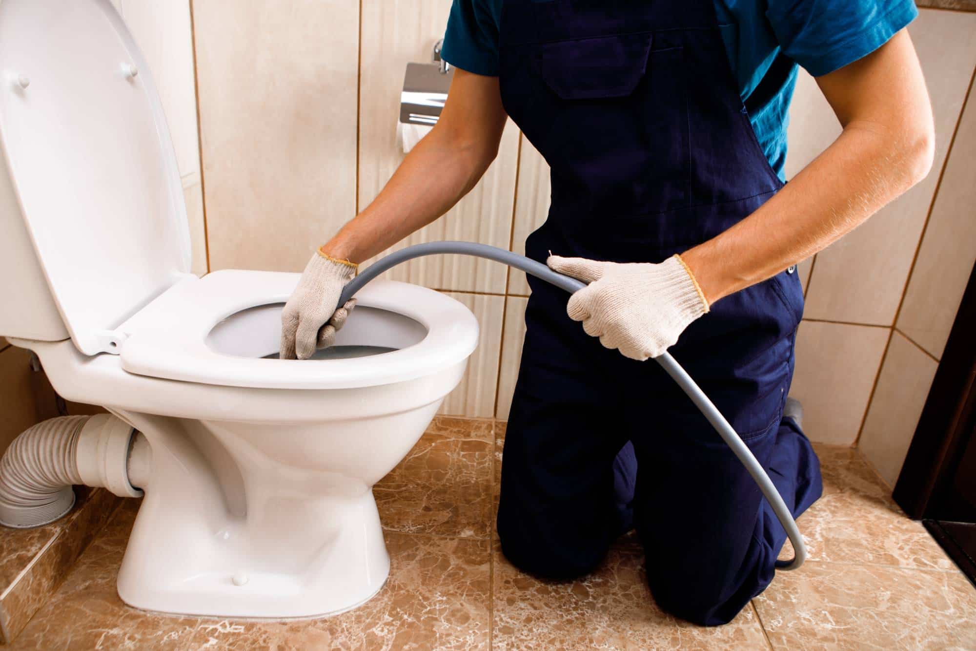 A person wearing gloves and overalls kneels on a tiled floor, using a flexible hose to clean or unclog a white toilet in a bathroom.
