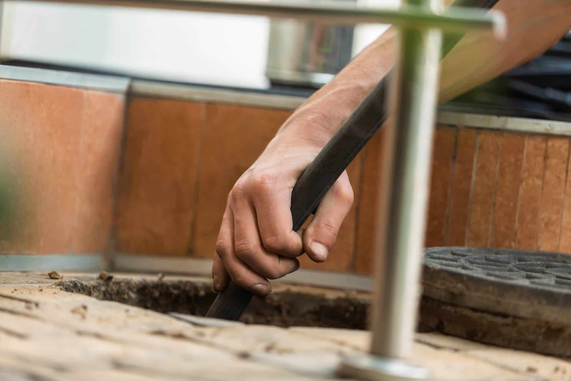 A close-up of a person’s hand holding a black hose or pipe, inserting it into an open drain or manhole, suggesting plumbing or drain cleaning work. Brown tiles and a metal cover are visible nearby.