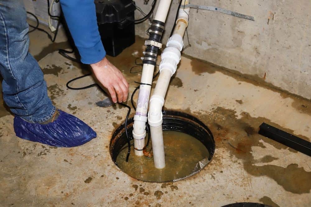 A plumber Chicago & Cook County, IL, wearing a blue shoe cover checks pipes connected to a sump pump in a basement, with water visible in the sump pit and water stains on the nearby concrete floor.