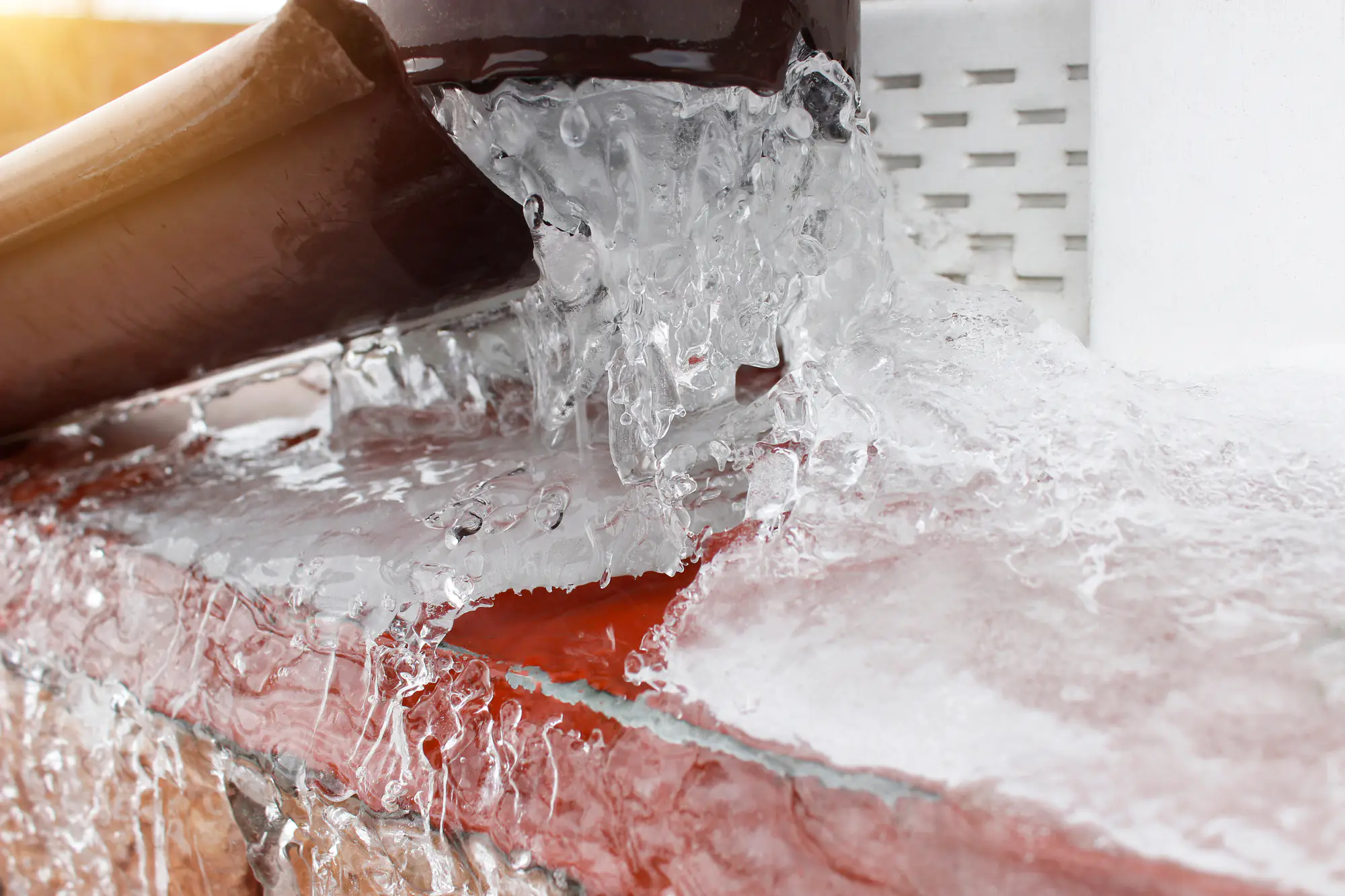 A brown gutter is overflowing with water that has frozen, creating icicles and a thick layer of ice over a red brick surface, illustrating winter freezing conditions.
