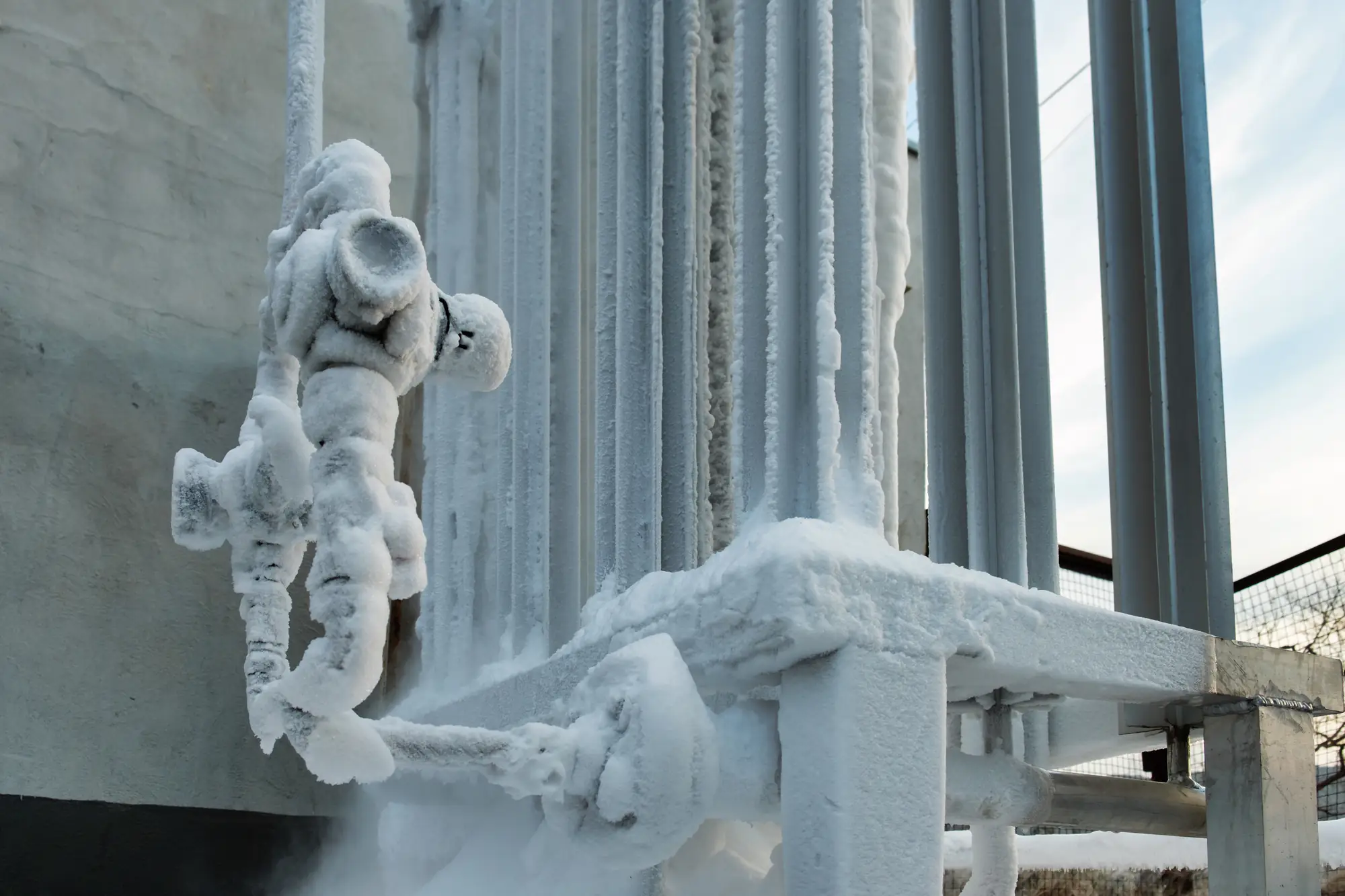 A metal pipe system and valve covered in thick white frost, likely from exposure to extreme cold or cryogenic materials, attached to an industrial structure outdoors.