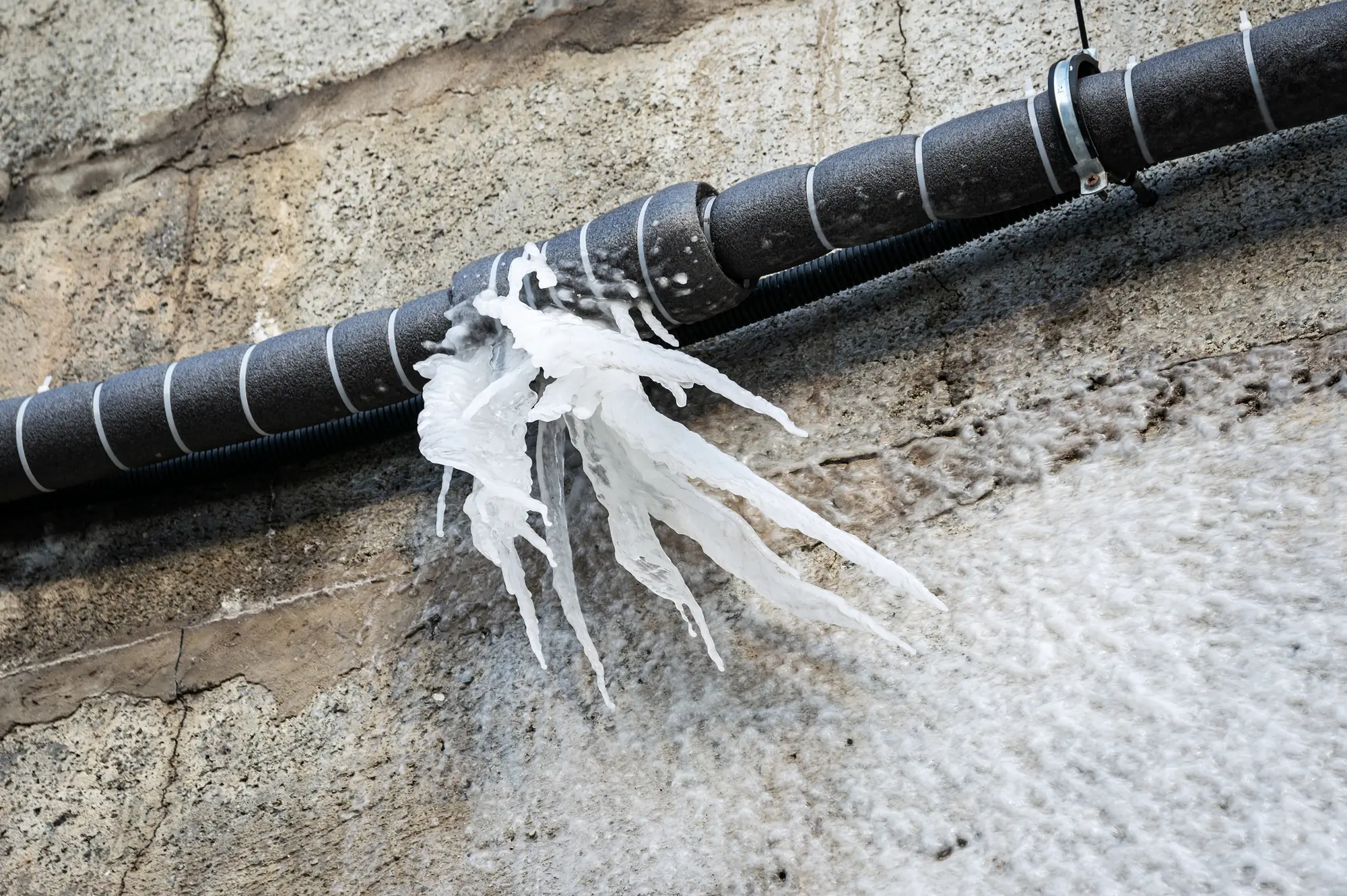 A black insulated pipe on a textured concrete wall with several long, jagged icicles hanging from a leak, creating dramatic frozen formations.
