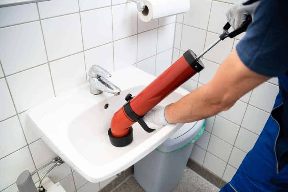 A person wearing gloves uses a red plumbing tool to unclog a white bathroom sink, with a trash bin and toilet paper visible in the background.