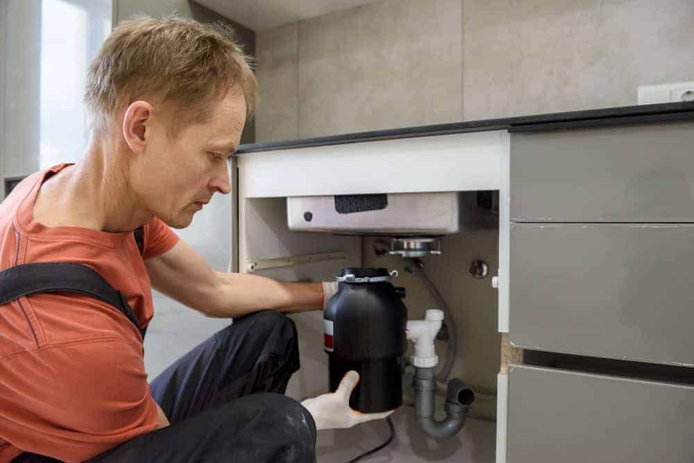 A person kneels on the floor in a kitchen while installing or repairing a garbage disposal unit under the sink, surrounded by pipes and gray cabinets.