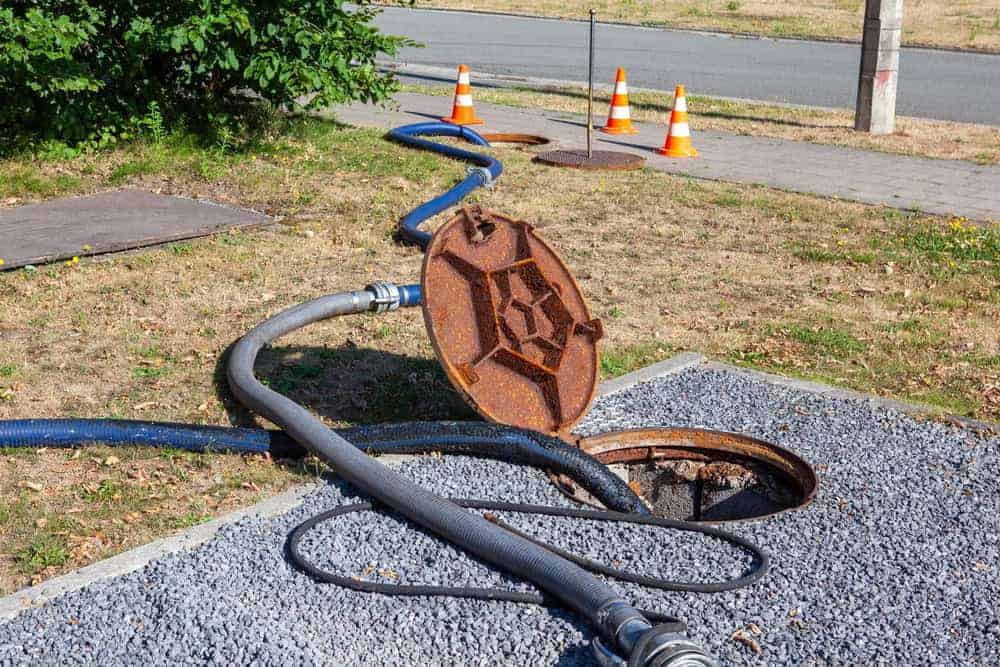 A rusty manhole cover is propped open with hoses leading inside. Nearby, another open manhole is surrounded by orange traffic cones. The scene is set near a road and grassy area.