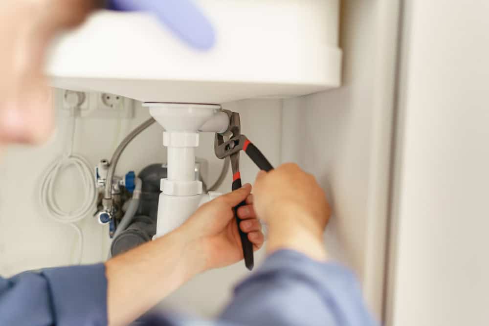 A person uses a wrench to tighten or loosen a pipe underneath a sink, performing plumbing work.