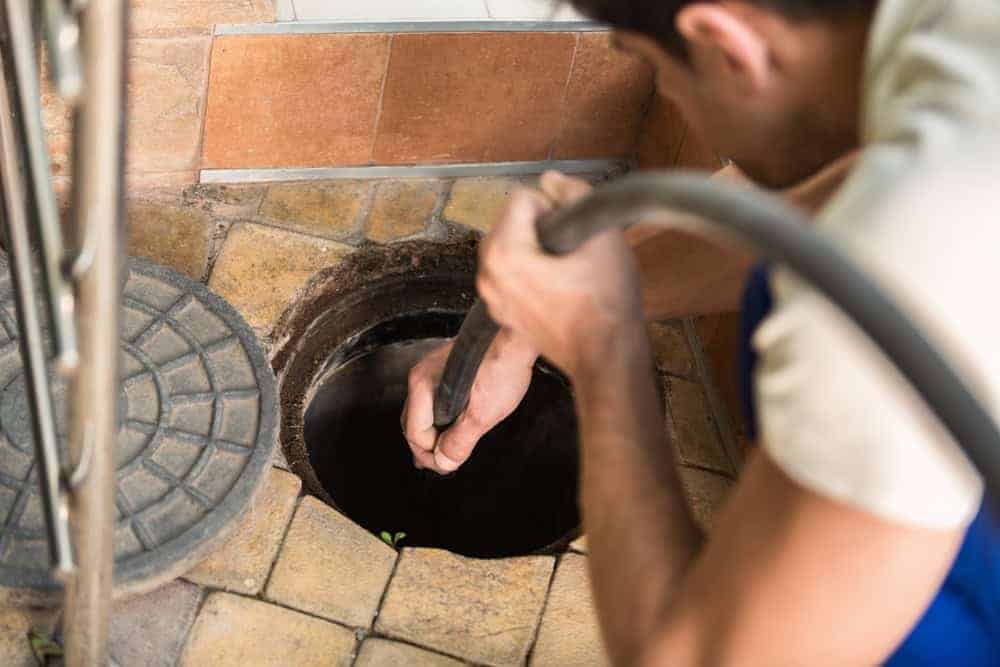 A person in a white shirt uses a hose to clean or unclog a circular drain or sewer opening set in a tiled floor. A round metal cover is placed nearby.