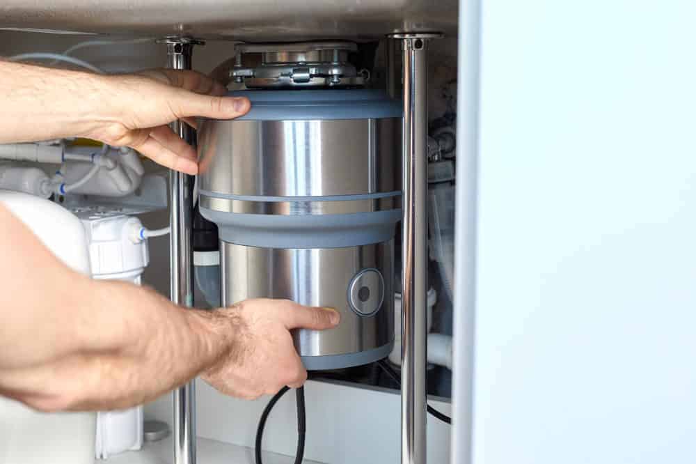 A person’s hands are installing or adjusting a stainless steel garbage disposal unit under a kitchen sink with visible pipes and plumbing components.