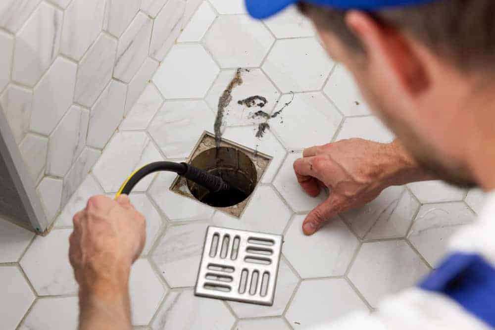 A plumber uses a tool to clear a clogged drain in a tiled bathroom floor, with the drain cover removed and placed nearby.