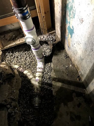 PVC plumbing pipes with connectors run vertically and branch horizontally through a gravel-filled trench in a basement with exposed framing and a worn, partially painted concrete wall.