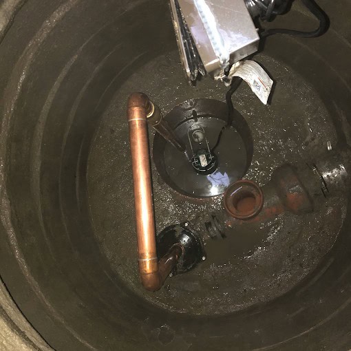 A view inside a sump pit showing copper pipes, a water pump, and electrical wiring. The interior appears wet and partially flooded, with reflections visible on the water surface.