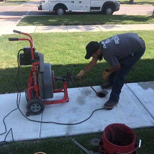 A worker wearing gloves and a cap is using a drain cleaning machine on a sidewalk near a grassy lawn, with tools and a red bucket nearby. A white utility truck is parked in the background.