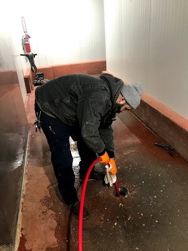 A person in a gray beanie and work clothes uses a red hose to clean a floor drain in a large, industrial space with concrete floors and white walls.