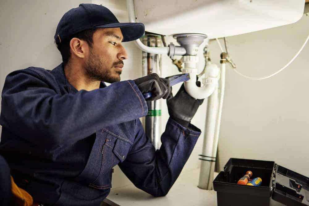 Plumber in a blue uniform repairs kitchen sink pipes in Chicago. Focus on maintenance and professional repair tools