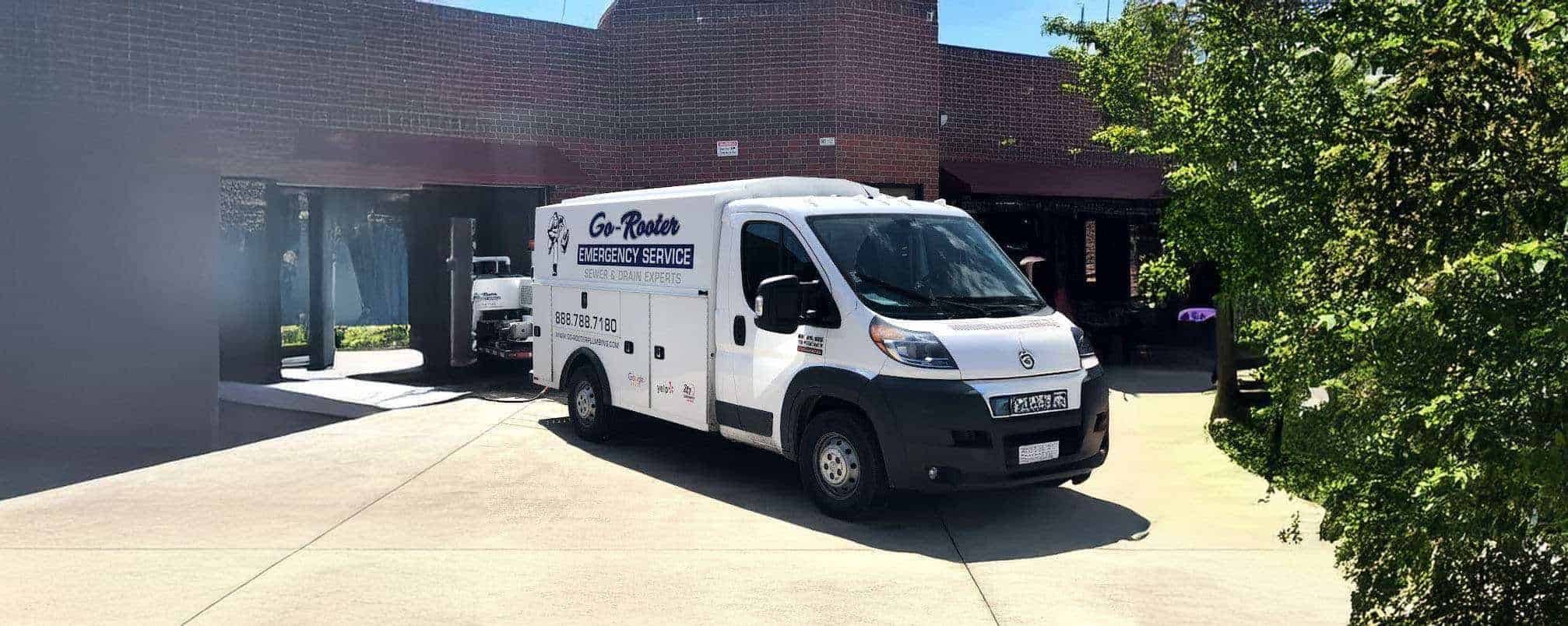 A white Go-Rooter emergency service van is parked on a driveway in front of a brick building, with trees providing some shade on a sunny day.
