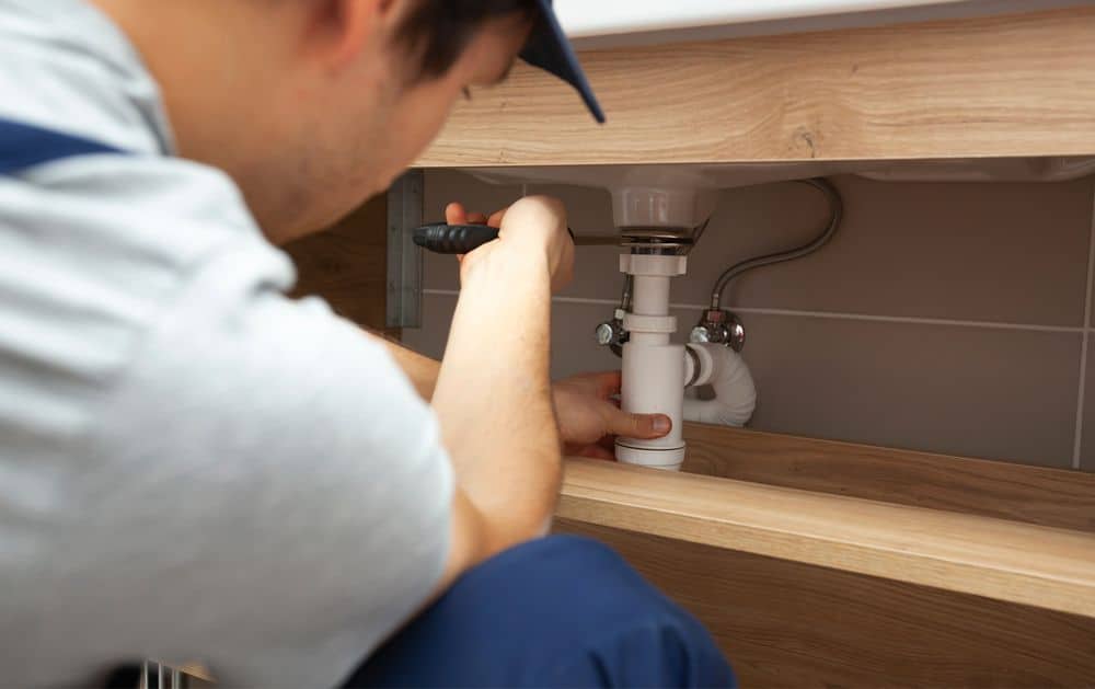 A plumber wearing a blue uniform and cap uses a screwdriver to repair a white plastic pipe under a bathroom sink. The plumber is focused on tightening connections in a wooden cabinet.
