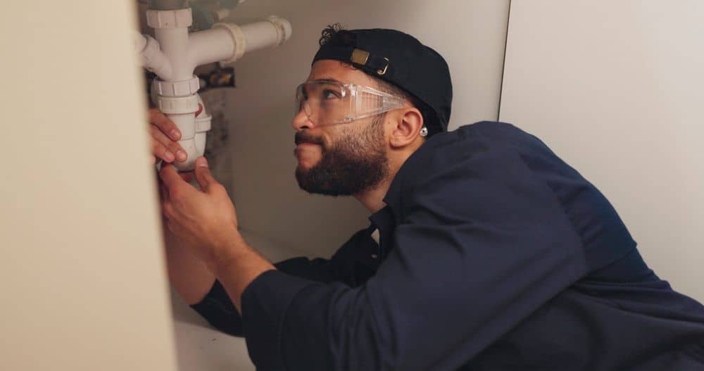 A person wearing safety glasses and a cap is lying under a sink, focused on fixing white PVC pipes with their hands.
