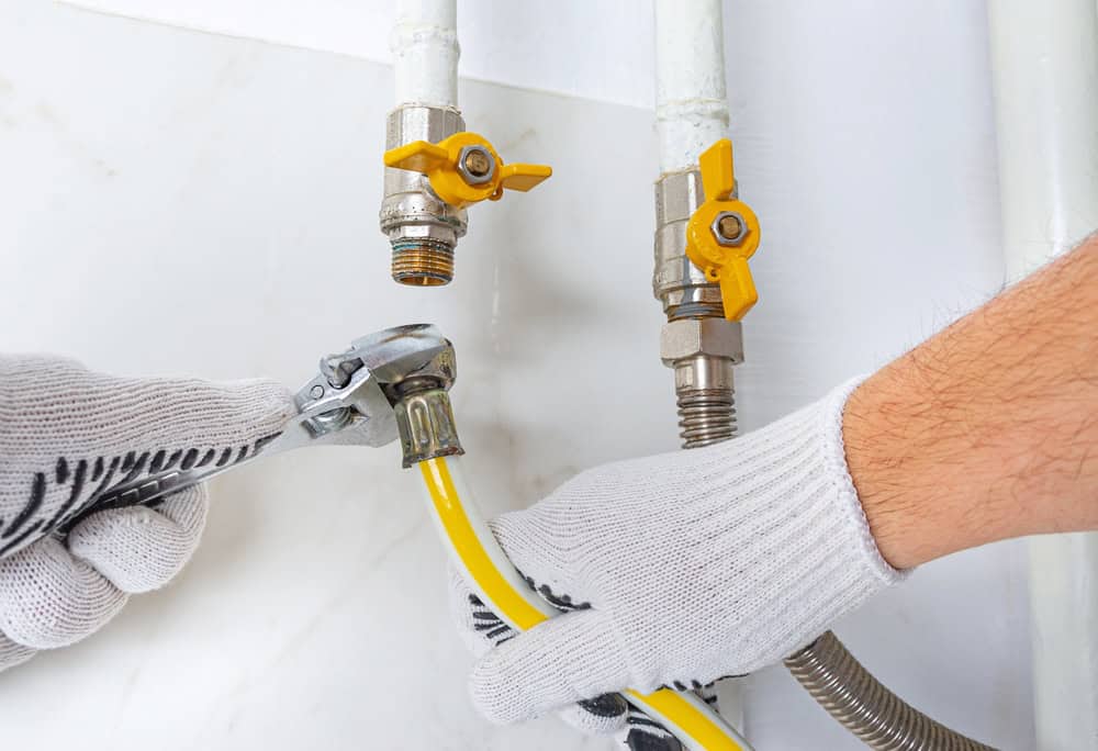 A person wearing white gloves uses a wrench to connect a yellow gas hose to a metal pipe with a shut-off valve. Two additional pipes with yellow valves are visible on a white wall.