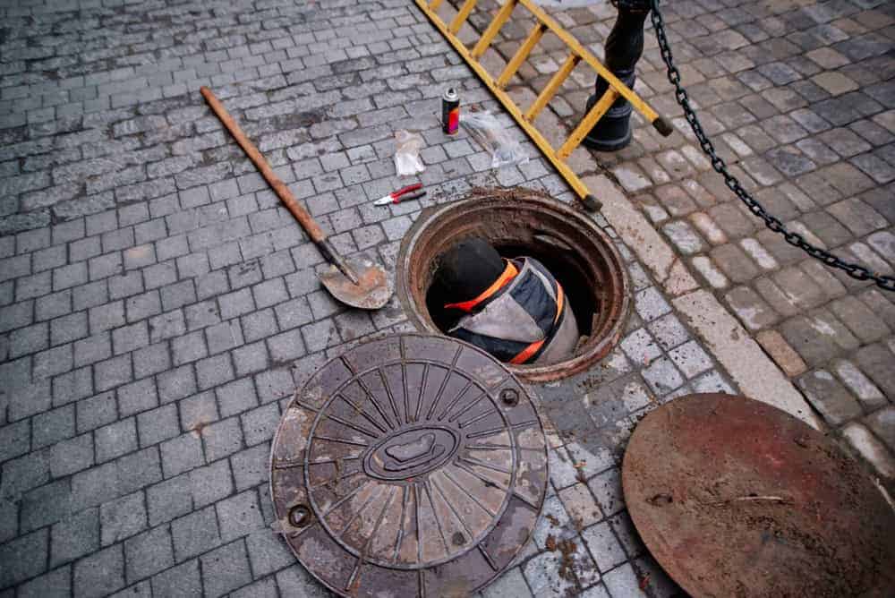 A worker wearing a safety vest enters an open manhole on a cobblestone street, surrounded by a shovel, ladder, tools, and two manhole covers.