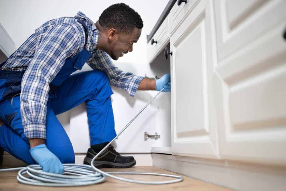 A plumber wearing blue overalls and gloves kneels on the kitchen floor, using a drain snake to unclog a pipe under a white cabinet.