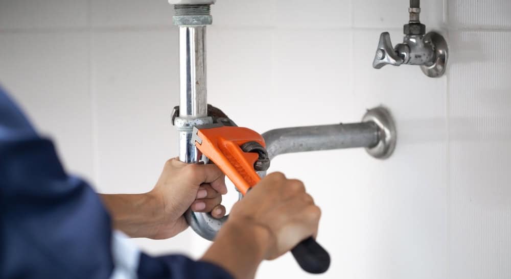 A person using an orange pipe wrench to tighten or fix a metal drain pipe under a sink against a white tiled wall.