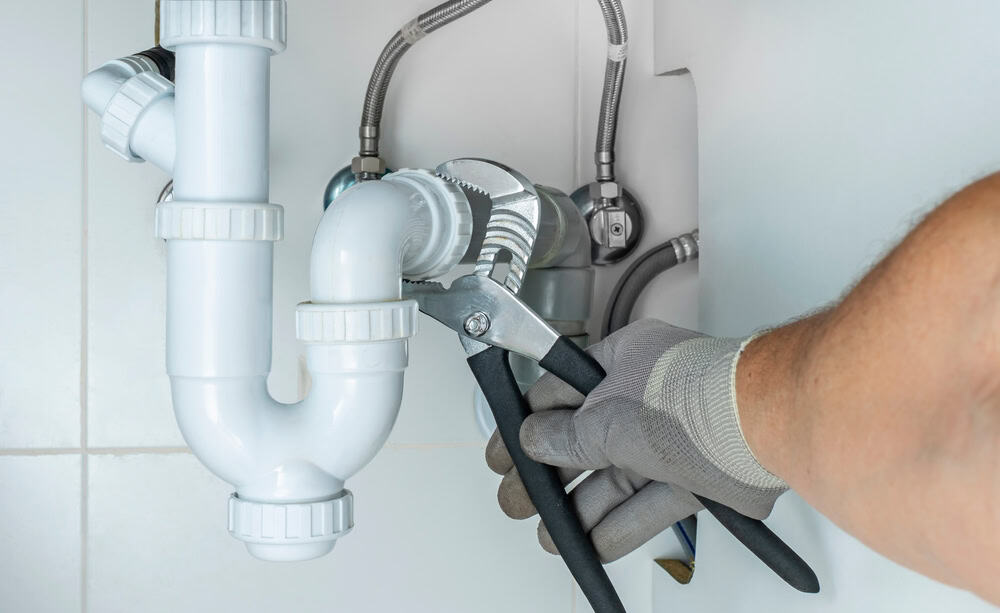 A person wearing a gray glove uses a wrench to tighten or loosen a pipe under a sink, surrounded by white plumbing and metal hoses against a tiled wall.